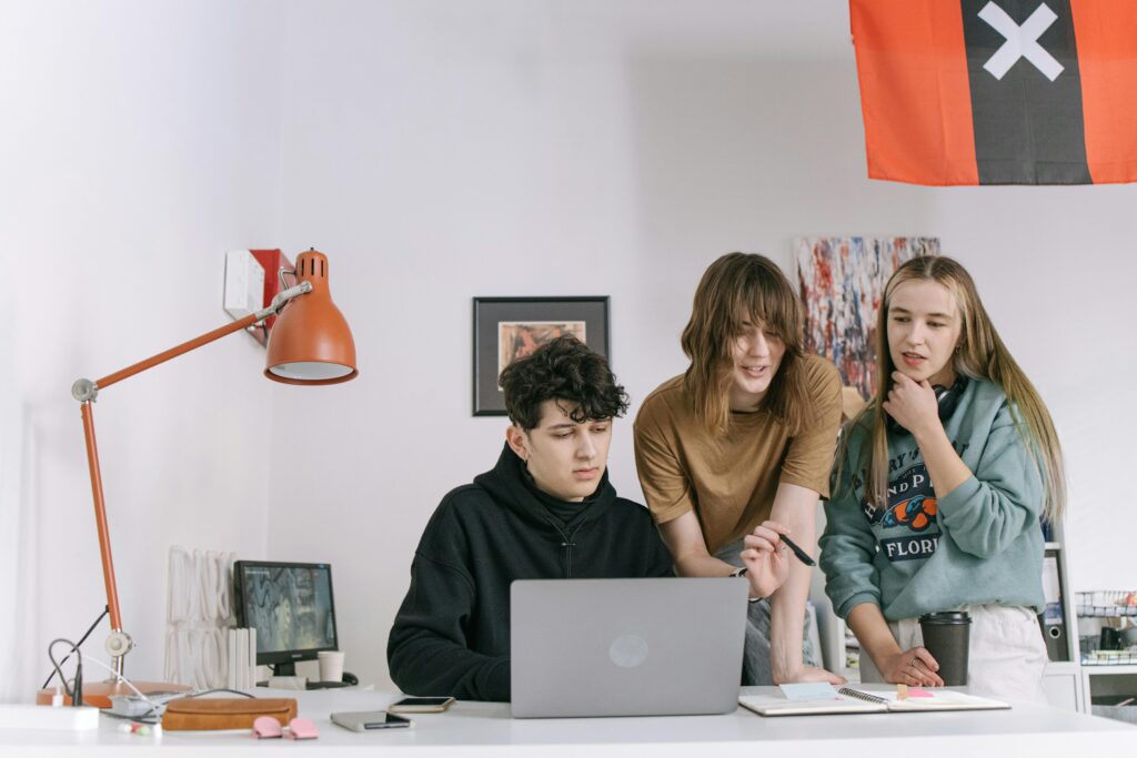A group of young adults working together in a modern office, focused on a laptop screen, illustrating teamwork and innovation.